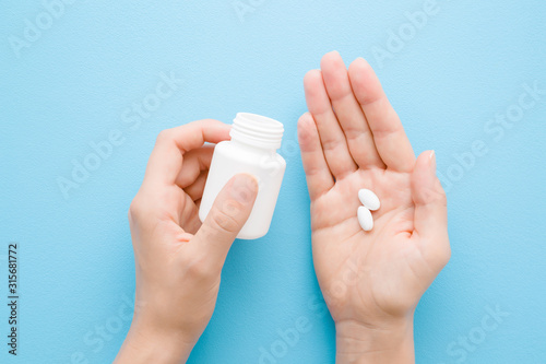 Young adult woman spilling out white pills from bottle in her hand. Receiving vitamins or medicaments. Point of view shot. Closeup. Pastel blue table background. Top down view.