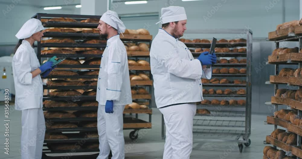 In a bakery industry main baker man taking photos from a new technology tablet of a baked fresh bread from shelves other two workers chatting on the background In a bakery industry main baker man taking photos from a new technology tablet of a baked fresh bread from shelves other two workers chatting on the background