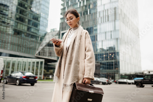 Cute Asian girl with glasses stands against the background of business skyscrapers and looks at the screen of her smartphone