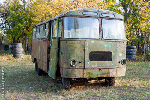 A green old rusty bus with traces of paint and with flat tires stands in a forest clearing among the trees. Shelter at the tactical paintball playground