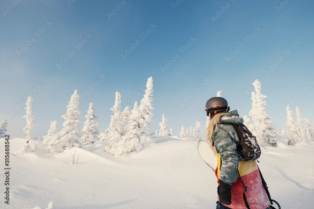 Female snowboarder wearing long dreadlocks and hoody holding a board, walking in snow-covered winter forest. Sheregesh, Siberia