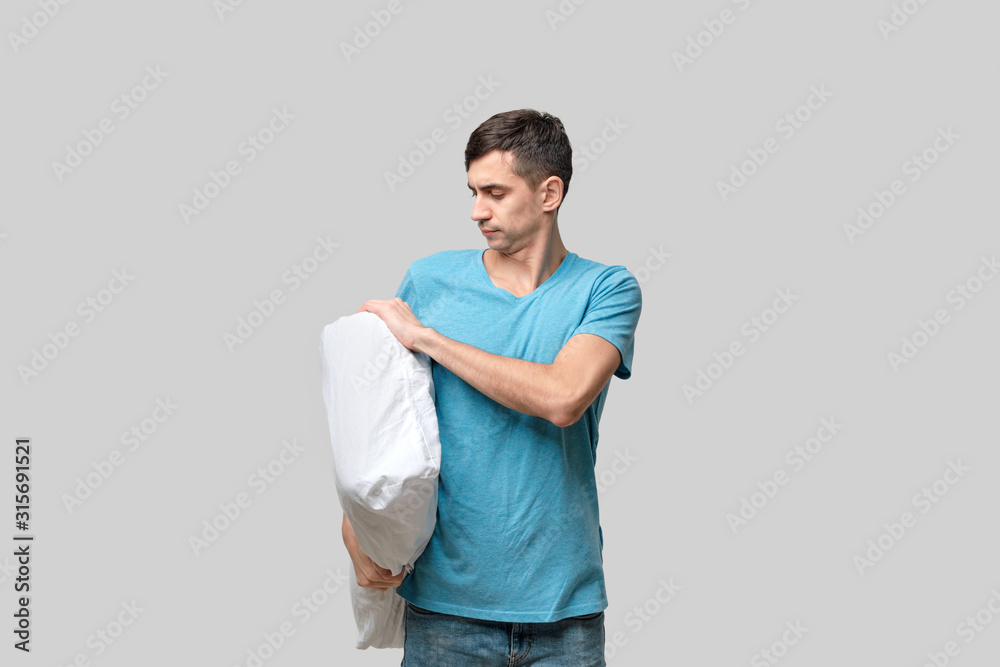 Tired brunet man in a blue tee holding white pillow isolated over grey background. Tired brunet man in a blue tee holding white pillow isolated over grey background.