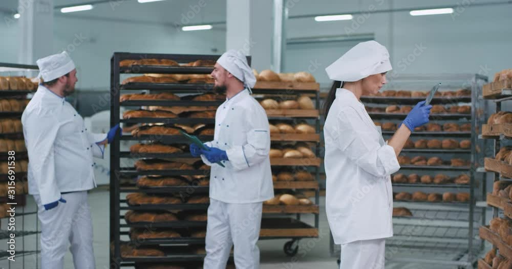 Bakery industry large video taking three bakers chatting with each other tone pretty lady baker in a white uniform taking photos of a fresh baked bread Bakery industry large video taking three bakers chatting with each other tone pretty lady baker in a white uniform taking photos of a fresh baked bread