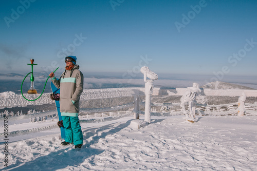 Snowboarder man relaxing and smoke a hookah or shisha, enjoying mountain view, winter frozen foggy day, snow and frost on the branch and table outdoor