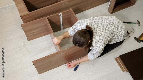 woman with dark hair sits on floor and assembles new prefabricated cabinet rotating tool to connect shelf parts upper view