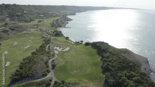 Aerial footage of Thracian Cliffs golf and beach resort. Beautiful landscape with green hills, meadows, golf course, pond, road, hotels, trees near the shore of Black sea, Bulgaria. Summer sunny day. photo