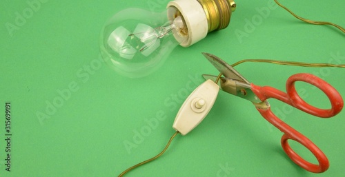 Ambiente, taglio degli sprechi energetici: forbici che tagliano il cavo di alimentazione di una vecchia lampadina a bulbo dotata porta lampada con isolante in ceramica, cavo e vecchio interruttore 