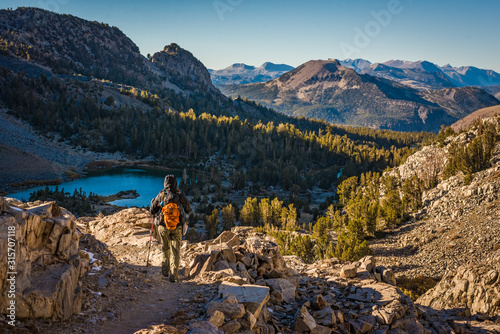 A hiker descends a granite trail to a forested lake in the mountains