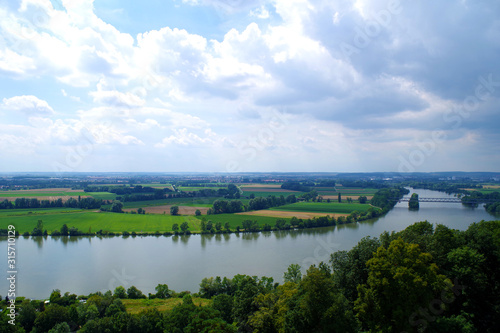 A landscape with the Danube river, viewed from Walhalla (Germany). Green and yellow fields dominate. Thick grove of trees in the foreground. Very distant towns can be seen. Dark clouds in the sky.