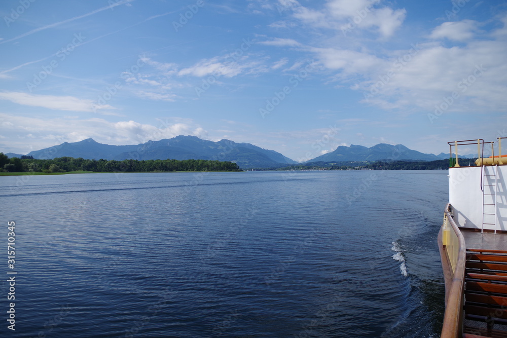 A view on the Chiemsee lake (Bavaria, Germany). There is an island and distant mountains visible on the horizon. A passenger ferry, in the foreground, is traveling the calm water. 