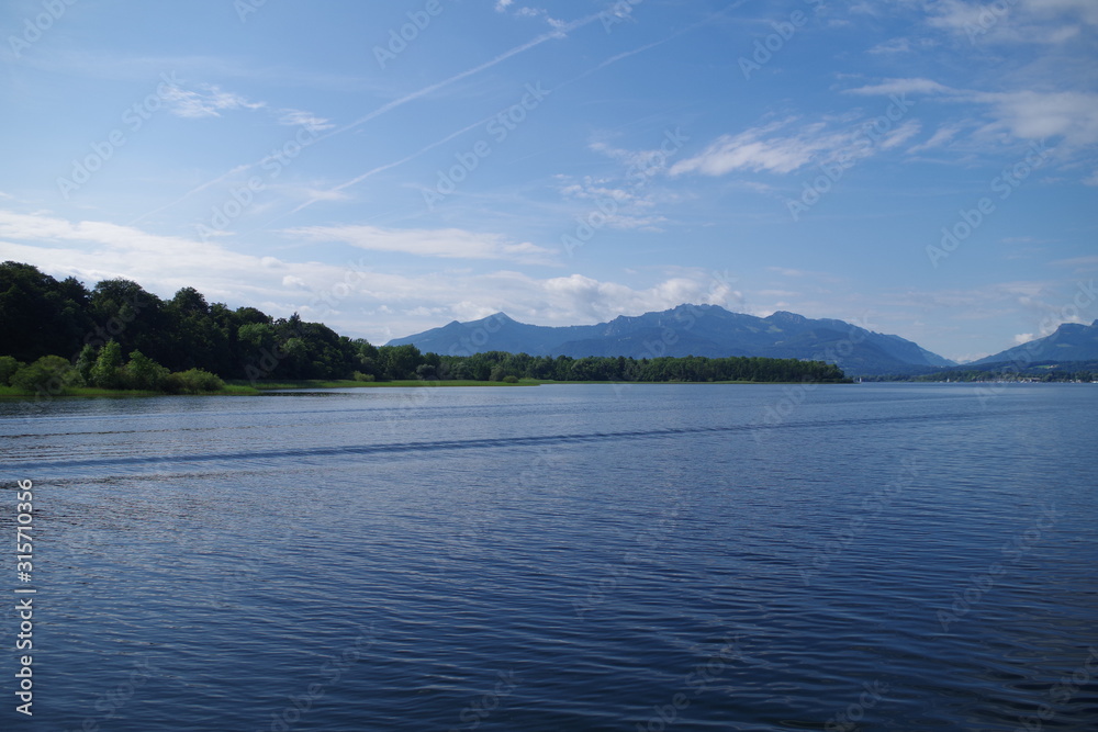 A view on the Chiemsee lake (Bavaria, Germany). In the distance, mountains can be seen, as well as forested islands. The sky is very blue, there are scattered clouds. Clam waters with slight ripples.