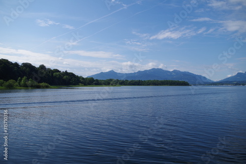 A view on the Chiemsee lake (Bavaria, Germany). In the distance, mountains can be seen, as well as forested islands. The sky is very blue, there are scattered clouds. Clam waters with slight ripples.