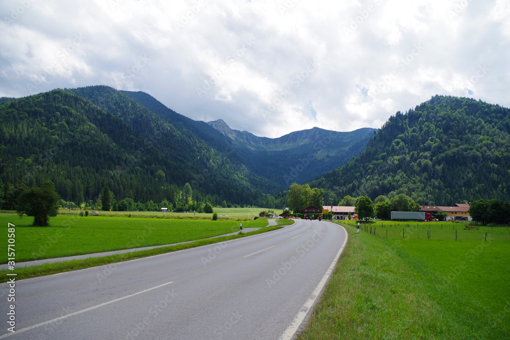 An empty asphalt road through the green fields leading towards the mountain village. In the distance, there are heavily forested green mountains and several buildings. Sky is very cloudy.