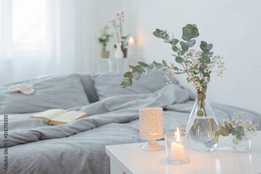 burning candles and eucalyptus in glass vase in white bedroom