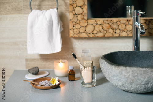 Natural bathroom accessories: bamboo toothbrush, make up remover in a glass container, coconut wax candle, organic bath salt, volcanic pumice and cotton towel standing near black stone sink.