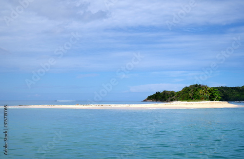 A shot of a very calm sea on a sunny day. There are few scattered clouds in the sky. On the horizon we can see a small island, overgrown with palm trees and sporting an inviting, sandy beach