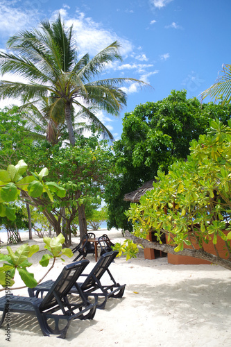 A small island in the Philippines archipelago. There are two lounge chairs standing on a sandy beach. A small hut stands between the palm trees. It's sunny and warm, white clouds on a blue sky. 