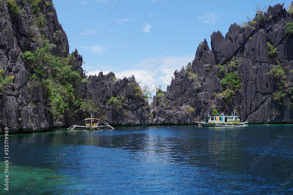 Big rocky island in the Philippines archipelago. Three paraw boats (double outrigger sail ships) are sailing near. The water of the ocean is calm and rippling slightly, it's a clear summer day.