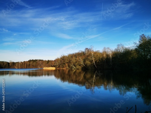 A quiet calm lake in January with different dark and light blue tones sky and trees that are reflected on the shore in the water and magnificent panorama with far-sightedness