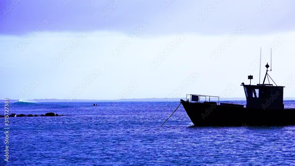 A fishing boat drifts on anchor in the evening in a calm bay with surfers in the distance
A fishing boat drifts on anchor in the evening in a calm bay with surfers in the distance