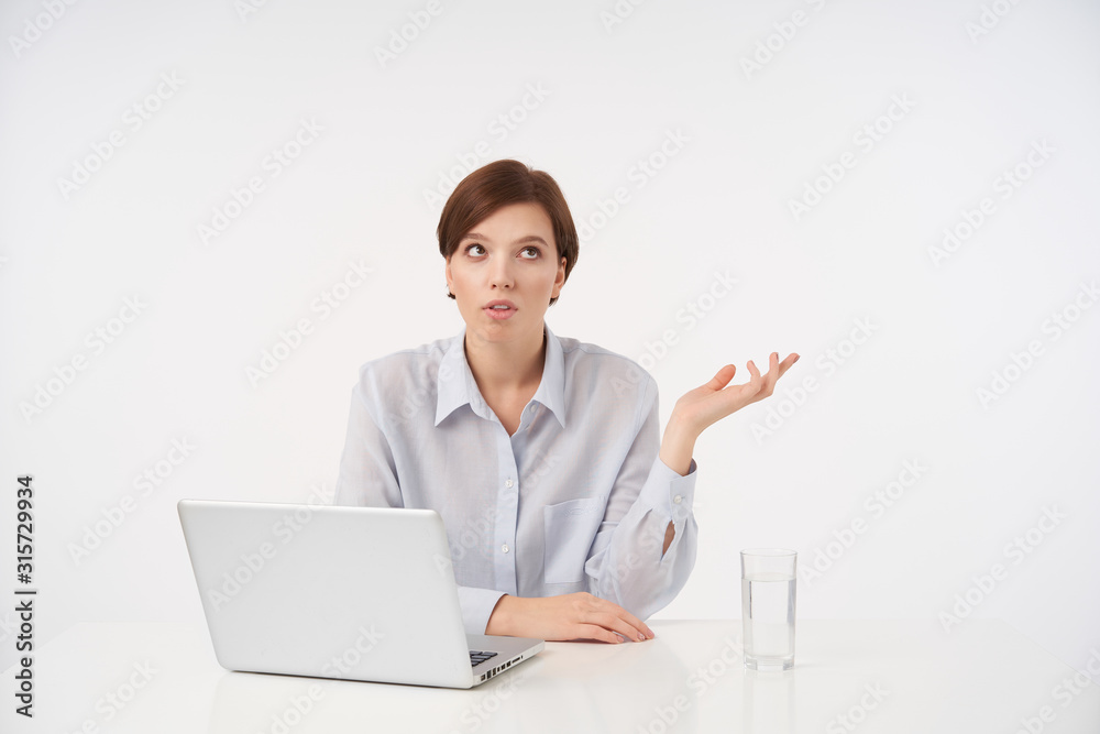 Displeased young brown-eyed short haired brunette woman sitting at table with laptop and looking aside with pout, raising up her palm while posing over white background Displeased young brown-eyed short haired brunette woman sitting at table with laptop and looking aside with pout, raising up her palm while posing over white background