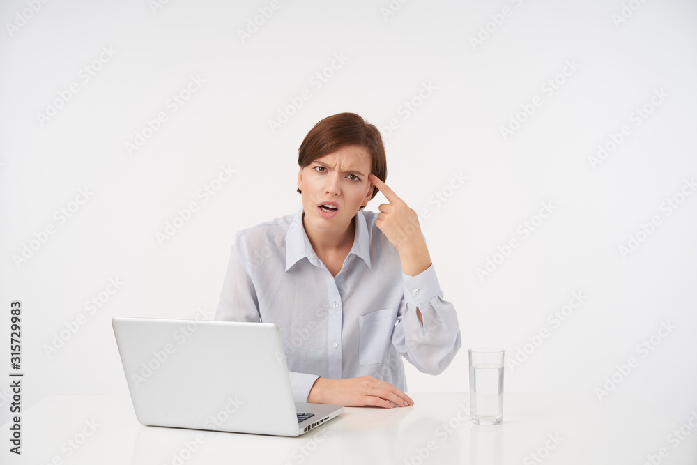 Indoor photo of young dissatisfied brown haired female folding gun with raised hand and keeping it on her temple, frowning eyebrows with pout while looking at camera Indoor photo of young dissatisfied brown haired female folding gun with raised hand and keeping it on her temple, frowning eyebrows with pout while looking at camera