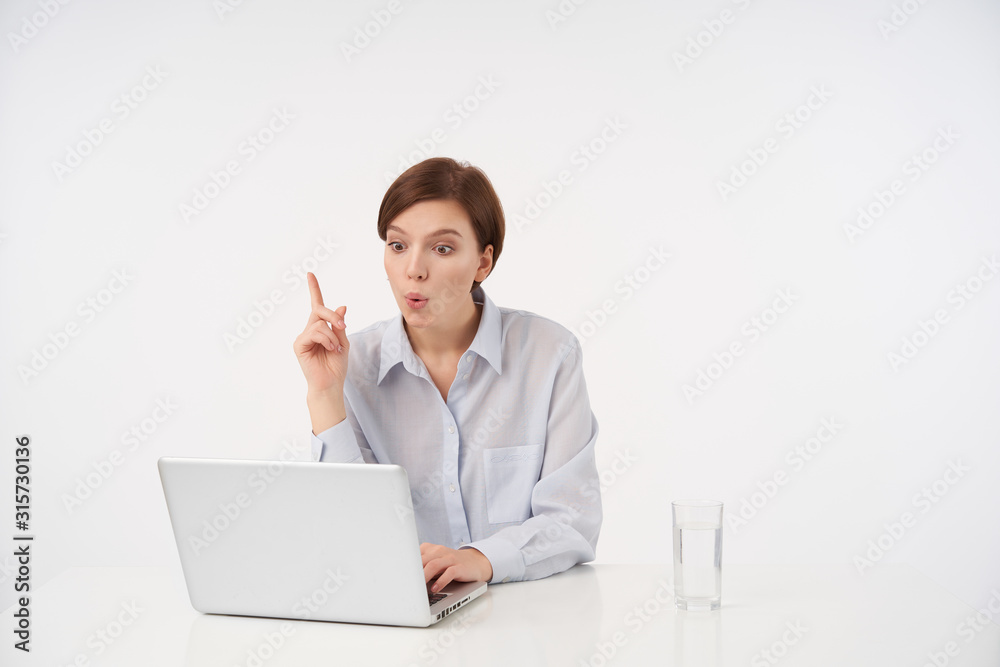 Indoor photo of attractive young short haired brunette female rounding excitedly her mouth and raising forefinger as she is having good idea, posing over white background Indoor photo of attractive young short haired brunette female rounding excitedly her mouth and raising forefinger as she is having good idea, posing over white background