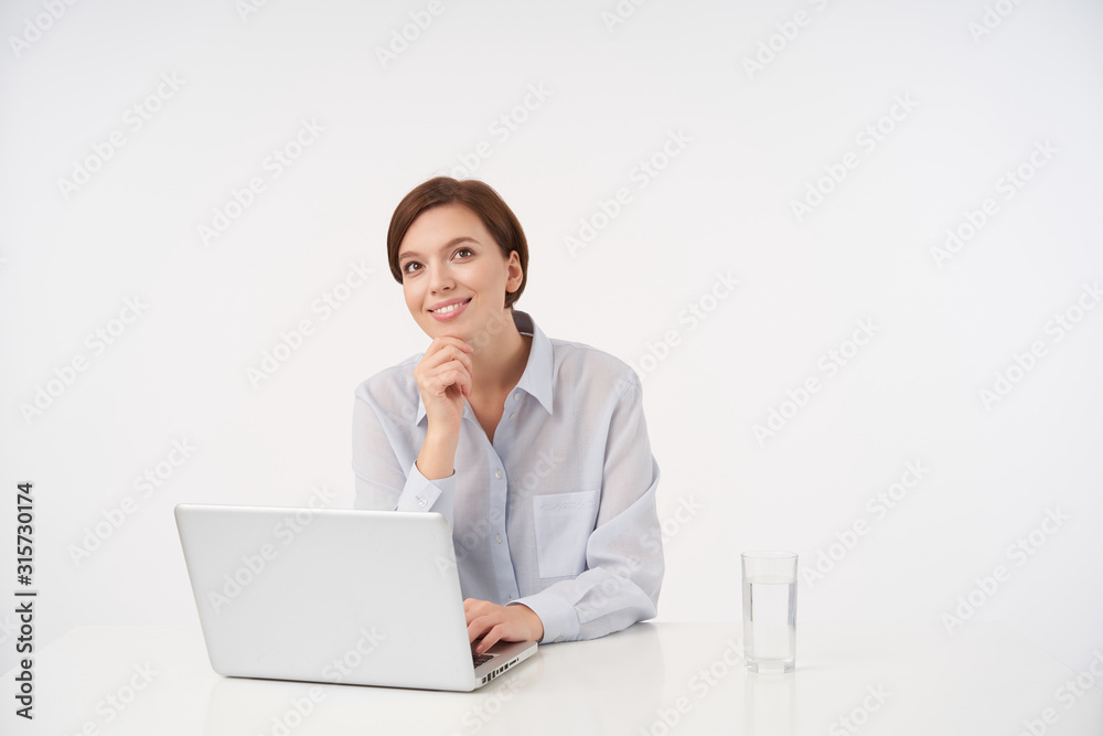 Positive young brown-eyed brunette female with casual hairstyle keepig raised hand on her chin and looking cheerfully aside with charming smile, sitting over white background Positive young brown-eyed brunette female with casual hairstyle keepig raised hand on her chin and looking cheerfully aside with charming smile, sitting over white background