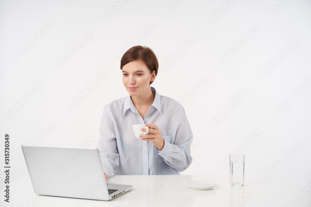 Indoor photo of pretty young brown haired woman with casual hairstyle drinking cup of coffee while working over white background with modern laptop and smiling positively Indoor photo of pretty young brown haired woman with casual hairstyle drinking cup of coffee while working over white background with modern laptop and smiling positively