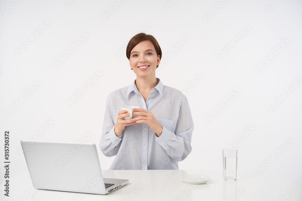 Beautiful young short haired brunette female with natural makeup looking positively at camera with charming smile, keeping cup of tea in raised hands, isolated over white background Beautiful young short haired brunette female with natural makeup looking positively at camera with charming smile, keeping cup of tea in raised hands, isolated over white background