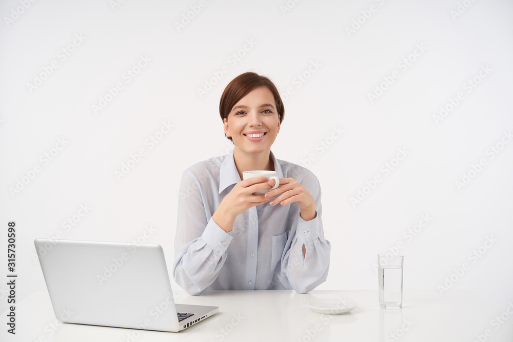 Charming young brown-eyed short haired lady holding cup of coffee in raised hands and looking cheerfully at camera with pleasant smile, working in modern office with laptop Charming young brown-eyed short haired lady holding cup of coffee in raised hands and looking cheerfully at camera with pleasant smile, working in modern office with laptop