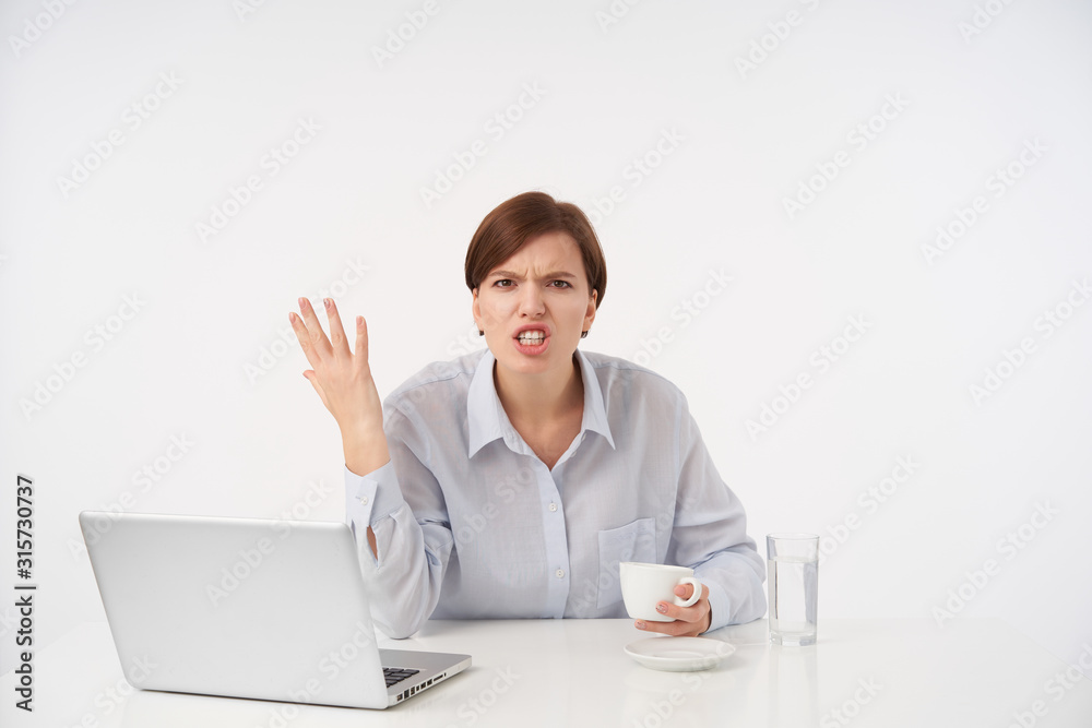 Irritated young brown haired woman with natural makeup raising emotionally her hand while looking angrily at camera, having stressed day, sitting over white background Irritated young brown haired woman with natural makeup raising emotionally her hand while looking angrily at camera, having stressed day, sitting over white background