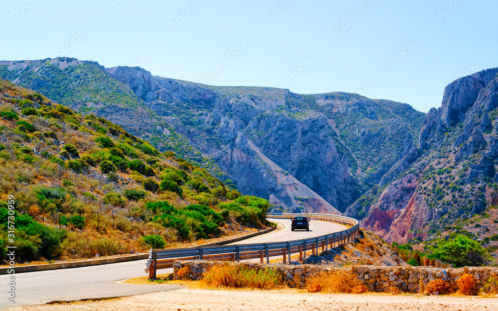Naklejka premium Car on the road at Buggerru in Sardinia Island in Italy summer. Transport driving on the highway of Europe. Holiday View on motorway. Cagliari province. Mountains on background. Mixed media.