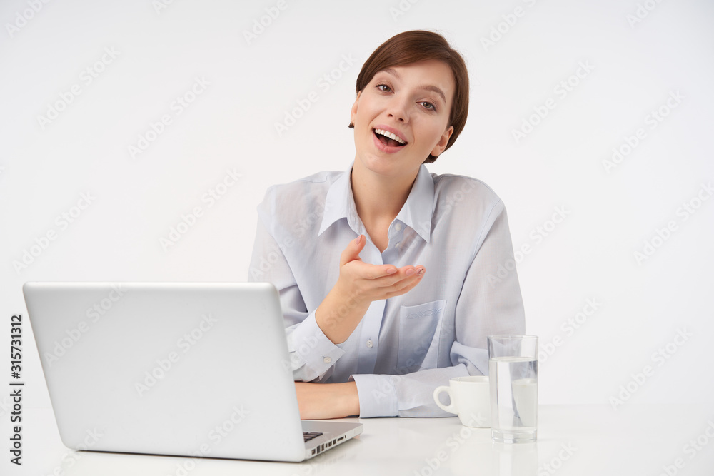 Indoor shot of joyful young brown-eyed short haired lady with casual hairstyle working in modern office with laptop, raising her palm and smiling cheerfully at camera Indoor shot of joyful young brown-eyed short haired lady with casual hairstyle working in modern office with laptop, raising her palm and smiling cheerfully at camera