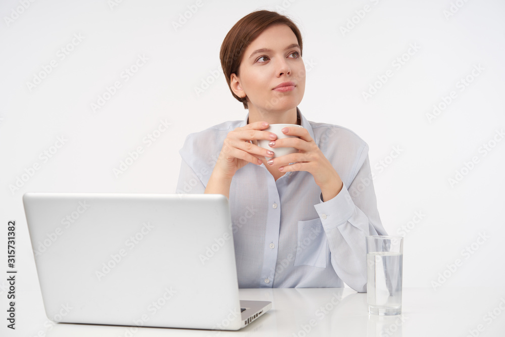 Good looking young brown haired female with natural makeup keeping cup of tea in raised hands while making break with her work, sitting over white background Good looking young brown haired female with natural makeup keeping cup of tea in raised hands while making break with her work, sitting over white background