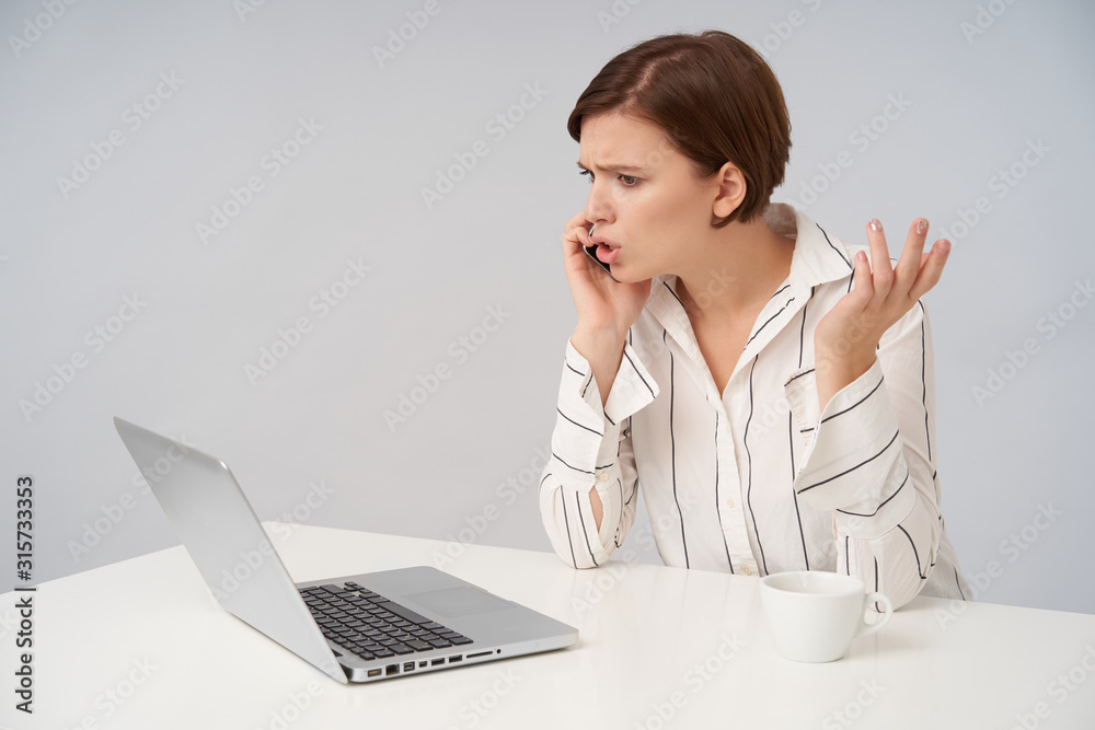 Confused young pretty short haired brunette female with casual hairstyle raising her palm while solving problems during phone call, frowning her face while looking in front of herself Confused young pretty short haired brunette female with casual hairstyle raising her palm while solving problems during phone call, frowning her face while looking in front of herself