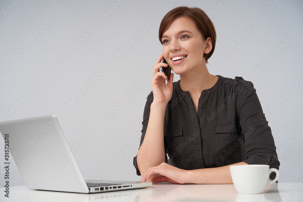 Cheerful young brown-eyed lovely brunette female with short trendy haircut smiling pleasantly while making call, sitting at table over white background with laptop and cup of coffee Cheerful young brown-eyed lovely brunette female with short trendy haircut smiling pleasantly while making call, sitting at table over white background with laptop and cup of coffee