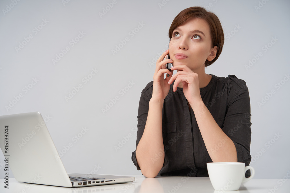 Indoor photo of young brown-eyed attractive brunette lady with natural makeup looking positively upwards with light smile while posing over white background in black shirt Indoor photo of young brown-eyed attractive brunette lady with natural makeup looking positively upwards with light smile while posing over white background in black shirt