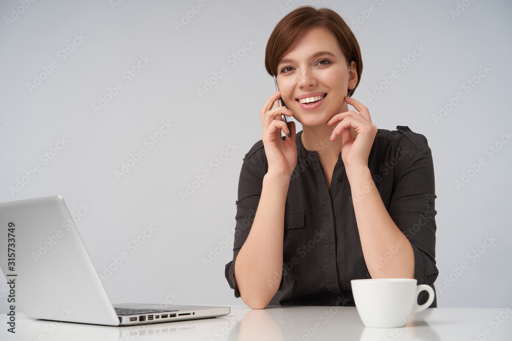 Beautiful young brown haired female with short trendy haircut looking positively at camera with charming smile while having pleasant phone conversation over white background Beautiful young brown haired female with short trendy haircut looking positively at camera with charming smile while having pleasant phone conversation over white background