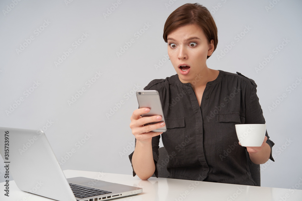 Shocked young attractive brown haired female with natural makeup holding mobile phone and looking at screen with wide eyes and mouth opened, sitting over white background Shocked young attractive brown haired female with natural makeup holding mobile phone and looking at screen with wide eyes and mouth opened, sitting over white background