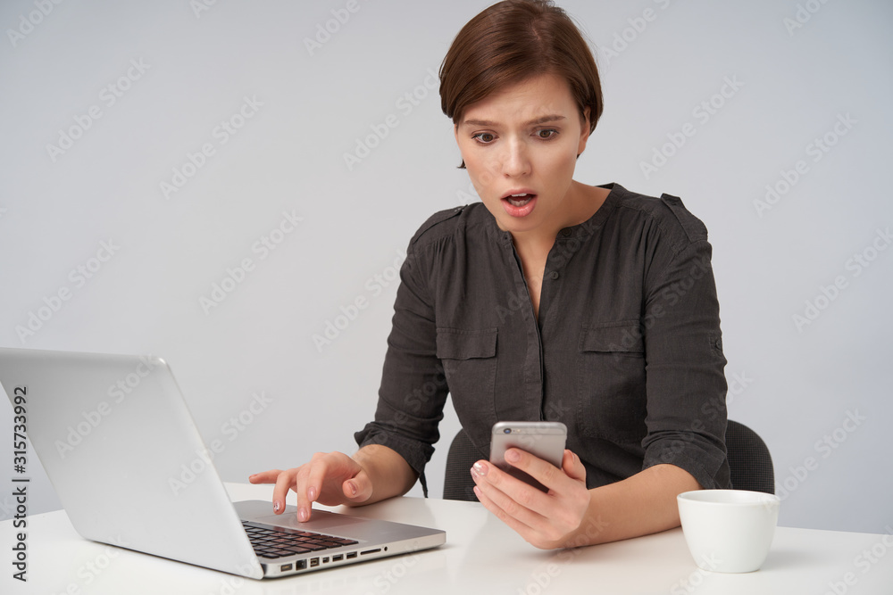 Indoor shot of young amazed short haired brunette lady with casual hairstyle looking confusedly at her smartphone while sitting at table over white background, dressed in black shirt Indoor shot of young amazed short haired brunette lady with casual hairstyle looking confusedly at her smartphone while sitting at table over white background, dressed in black shirt