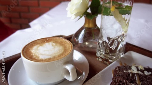 Breakfast: chocolate brownie, cappuccino, a glass of water with lemon and flowers on the table