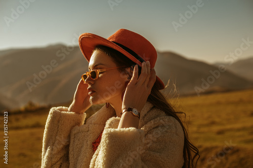 Outdoor close up fashion portrait of young beautiful confident brunette woman wearing stylish orange hat, sunglasses, wrist watch, faux fur coat, posing in mountain landscape. Copy, empty space 