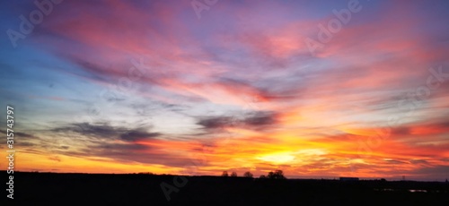 A fantastic sunset with red yellow orange and purple tones with dramatic cloud formation over green meadows with bare trees in January on the island of Rügen