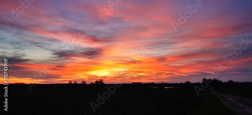 A fantastic sunset with red yellow orange and purple tones with dramatic cloud formation over green meadows with bare trees in January on the island of Rügen