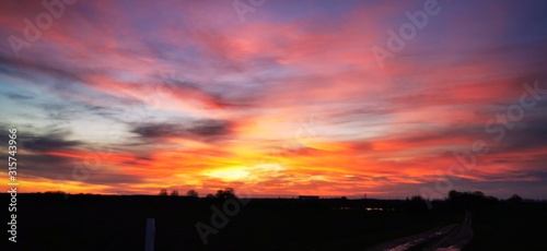A fantastic sunset with red yellow orange and purple tones with dramatic cloud formation over green meadows with bare trees in January on the island of Rügen