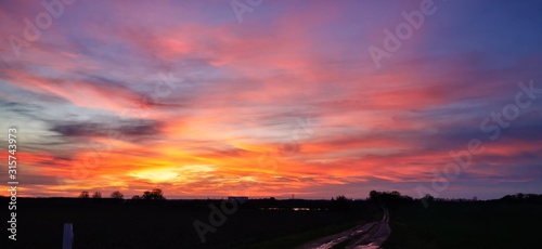 A fantastic sunset with red yellow orange and purple tones with dramatic cloud formation over green meadows with bare trees in January on the island of Rügen