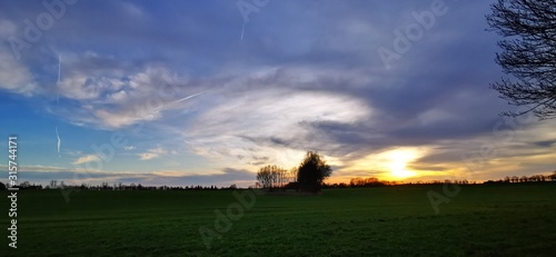 A fantastic sunset with red yellow orange and purple tones with dramatic cloud formation over green meadows with bare trees in January on the island of Rügen