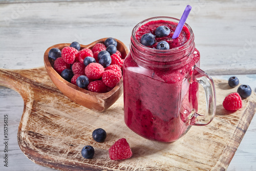 Fresh smoothies in a glass jar with ripe raspberries and blueberries in a wooden heart-shaped bowl on rustic cutting board. Healthy food concept. Dessert for valentines day