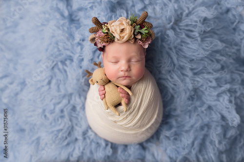 the first photo session of a newborn. newborn baby girl pose cocoon with a headband with horns and a deer toy on a blue background